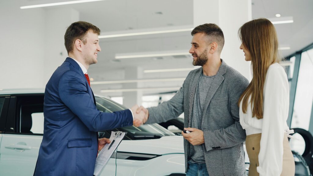 Friendly handshake between a car salesman and customers in a modern car showroom.